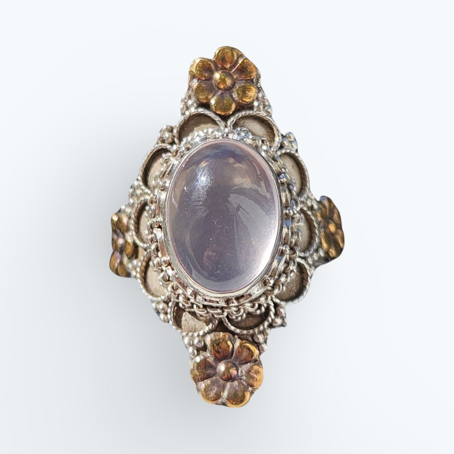 An ornate vintage-style silver ring featuring a large oval Rose quartz stone in the center, surrounded by intricate filigree detailing and small gold-toned floral accents 
